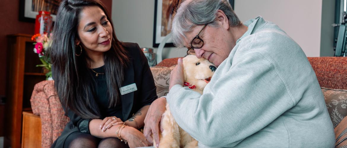Care home manager comforting a dementia resident with a robotic therapy dog in a lounge